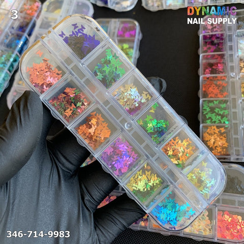 A gloved hand holds a clear plastic container with multiple compartments, each filled with vibrant holographic star-shaped nail design glitters perfect for creating glitter nail art. Similar containers can be seen in the background. The text reads Dynamic Nail Supply and 346-714-9983 in white.