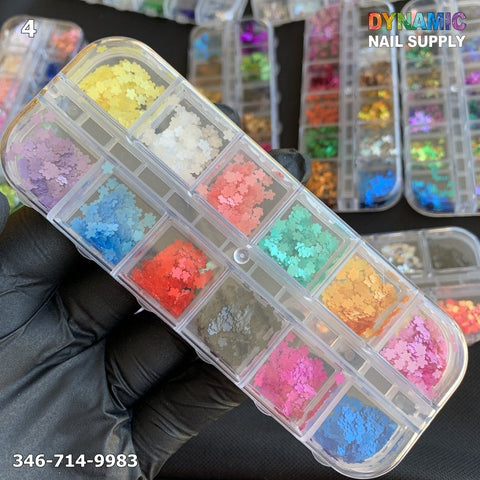 A hand wearing a black glove holds a transparent container featuring twelve compartments, each filled with vibrant, star-shaped glitter ideal for nail design. The glitters come in various shades including yellow, white, red, blue, green, and more. The information "Dynamic Nail Supply" and the number 346-714-9983 are visible in the image.