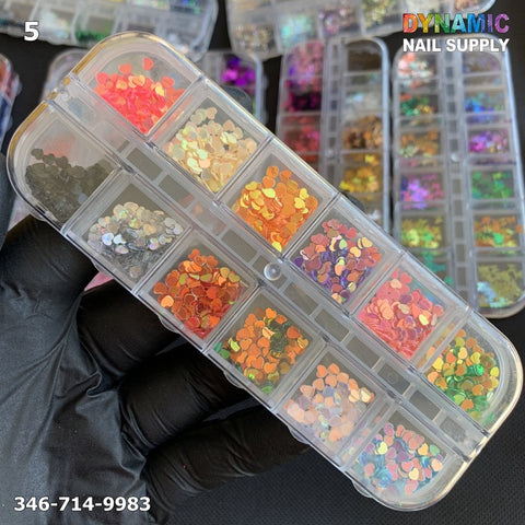 A gloved hand holds a rectangular container filled with twelve compartments of small, colorful, round NAIL DESIGN GLITTERs in various shades. The background features additional similar containers. The top right displays the logo "Dynamic Nail Supply," while the bottom left includes a phone number for all your nail design needs.