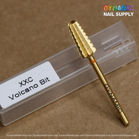A close-up of the high-quality, gold-colored 2XC Volcano Bit made from tungsten carbide. It is displayed on a transparent plastic case with "DYNAMIC" etched on its side. The packaging prominently features the brand name "Dynamic Nail Supply" in colorful letters at the top right corner, set against a wooden surface.