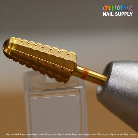 Positioned horizontally against a soft brown gradient background, a close-up view of a gold-colored 2XC Volcano Bit from Dynamic Nail Supply showcases its cylindrical shape and ridged edges, crafted from tungsten carbide. This superior quality drill bit is attached to a metallic handle, with the Dynamic Nail Supply logo displayed in vibrant colors in the top right corner.