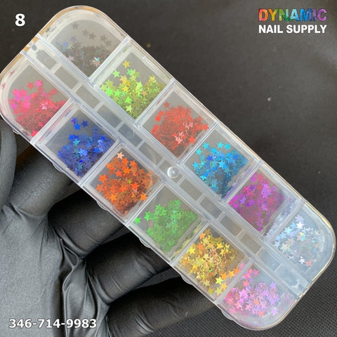 A clear plastic container with twelve compartments from Dynamic Nail Supply holds vibrant, star-shaped nail design glitter, organized by color. The shades include red, green, blue, orange, pink, and yellow. A hand in a black glove presents the container labeled "NAIL DESIGN GLITTER 8" by Dynamic Nail Supply. The top reads 346-714-9983.