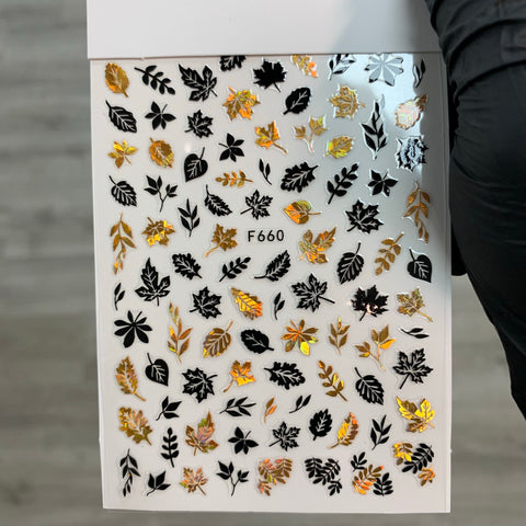 A sheet of Dynamic Nail Supply's FALL Nail Designs Stickers for Fall Season 2021, labeled F660, features captivating nail art decals. These stickers showcase black and gold leaves in various shapes and sizes with intricate detailing on a glossy finish, set against a plain background. The sheet is held by someone wearing a black glove.