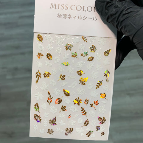 A hand in a black glove holds a sheet of FALL Nail Designs Stickers for Fall Season 2021 by Dynamic Nail Supply, ideal for Fall Nail Designs. The stickers feature leaf shapes in metallic shades of orange, green, and gold, perfectly encapsulating the essence of the season against a soft gray background.