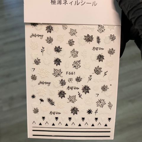 A sheet of FALL Nail Designs Stickers for Fall Season 2021 by Dynamic Nail Supply features black and white designs including leaves, the words Autumn and Harmony, as well as geometric shapes like triangles and stripes. Identified as F661, it is gracefully displayed by a gloved hand against a blurred background.