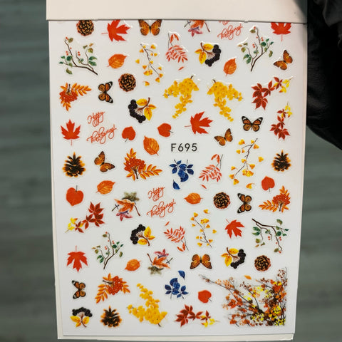 The F695 Dynamic Nail Supply FALL Nail Designs Stickers for Fall Season 2021 features trendy autumn-themed designs with orange and red leaves, pinecones, butterflies, and branches. "Happy Autumn" is playfully scattered amongst the stickers on a light background, making it perfect for celebrating the fall season!