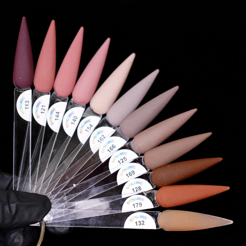 A fan of long, pointed nail polish swatches on clear sticks, held by a gloved hand against a black background. The swatches exhibit various earth tones with Dynamic Nail Supply's 2024 Fall Acrylic Powder Collection (part 1) Brown and Dark shades, ranging from deep burgundy to light beige to rich brown. Each is labeled with a small circular sticker indicating color numbers.