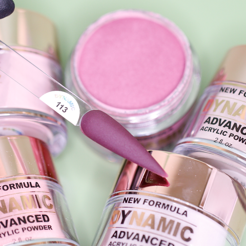 Close-up of several containers from the Dynamic Nail Supply's 2024 Fall Acrylic Powder Collection (Part 1), featuring brown and dark shades. The focus is on a deep pink shade labeled 113 on a swatch stick. The containers are arranged on a pale green surface, with shimmering reflections visible on their shiny silver lids and labels.