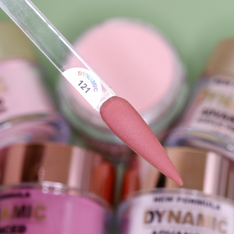 Close-up of a nail sample in a translucent holder, painted in a matte dusty rose shade. A label reads DYNAMIC 121. In the background, slightly blurred, are jars with metallic lids containing the "2024 Fall Acrylic Powder Collection (part 1) Brown and Dark shades" by Dynamic Nail Supply, perfect for creating long-lasting nails with pink and rose tones.