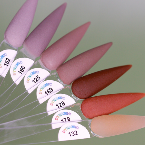 A close-up showcases eight pointed nail polish swatches from the Dynamic Nail Supply's 2024 Fall Acrylic Powder Collection (part 1) Brown and Dark shades, fanned out beautifully. The colors, ranging from light lavender to peachy brown, are crafted with long-lasting acrylic powder and feature labeled numbers beneath the DYNAMIC logo. The blurred green background adds a touch of fall elegance.