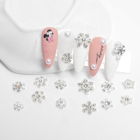 Displayed on a plastic stand by Dynamic Nail Supply is a set of nail art designs. Two nails showcase pink hues adorned with pearls and cartoon character decals, while three feature white coloring with snowflake embellishments and pearls. Below, additional silver snowflake charms from the "24pcs Snowflake Charms for Winter and Christmas Nails Art Design" collection are scattered, making it ideal for experimenting with various nail shapes.