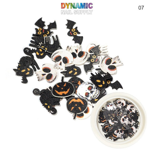 Halloween Sequins for Nail Art Design by Dynamic Nail Supply are scattered across a white background. These vibrant decorations include black bats, white skulls with red eyes, grinning orange pumpkins, ghosts, and witch hats. In the bottom right corner, a small round container filled with similar sequins is visible.