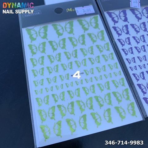 On a black surface lies a sheet of holographic metallic neon green butterfly nail stickers, featuring butterflies in different sizes, priced at $3.00 in the top right corner. In the background, you can see the edge of a second holographic sheet showcasing purple butterflies with the brand name Dynamic Nail Supply and a phone number visible.