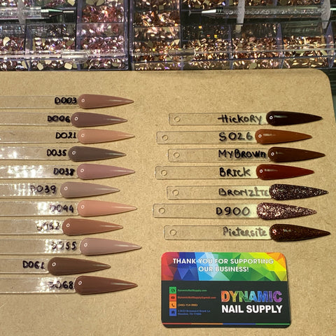 A display of nail polish samples on a table showcases pointed acrylic tips featuring a range of brown shades, labeled with names such as Hickory, MyBrown, and Piedrasite. Each sample is accompanied by the vibrant business card for Dynamic Nail Supply, renowned for their Dynamic 2-in-1 powder - Nude Collection 05 - Part 2, specializing in dipping nails and acrylic powder.