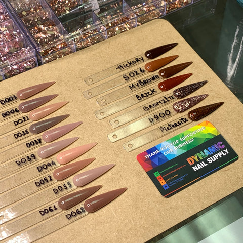 A wooden board adorned with diverse nail polish swatches, featuring labels such as Hickory and Picatrix, showcases a range of shades including nude, brown, and glittery dark. Nearby lies a vibrant Dynamic Nail Supply card next to the Dynamic 2-in-1 powder from their Nude Collection 05 - Part 2 line, alongside an assortment of dipping nails and nail powder for the perfect manicure finish.