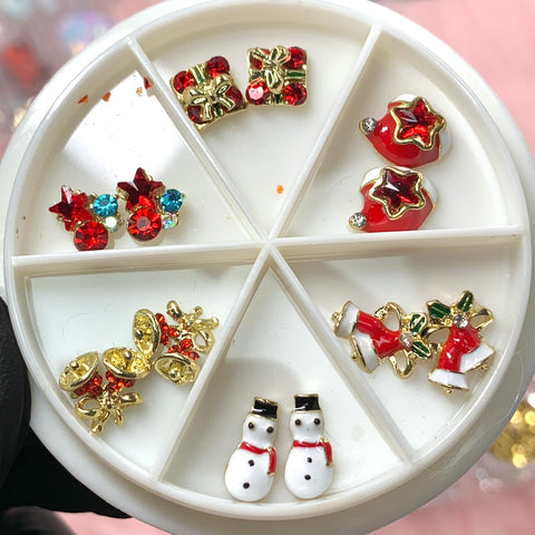 The 12 pcs Christmas Nail Design Charms by Dynamic Nail Supply includes a round, divided container filled with holiday-themed nail decorations ideal for DIY crafts. The set features sections with intricate, shiny charms such as red and gold gift boxes, star-shaped designs, Santa hat motifs, gold and red reindeer, Santa suits, and snowman figures. Each piece is small and exquisitely detailed to create unique nail art.