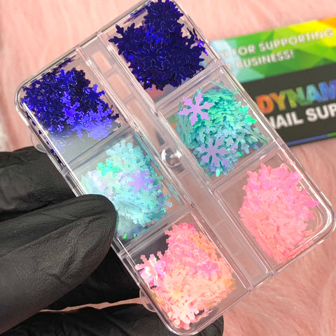 A hand adorned with a black glove holds a clear plastic container featuring six compartments, each filled to the brim with the Special Iridescent Snowflakes Sequin Glitter for Christmas Nail Art design by Dynamic Nail Supply. The glitters shimmer in shades of blue, turquoise, silver, pink, and iridescent tones. In the background, a colorful business card is slightly blurred.