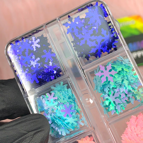 A gloved hand showcases the "Special Iridescent Snowflakes Sequin Glitter for Christmas Nail Art Design" by Dynamic Nail Supply, with a clear plastic organizer featuring six compartments of shimmering snowflake glitter and metallic nail art sequins in mesmerizing shades of blue, purple, teal, and pink against a blurred backdrop with a hint of pink and a colorful logo.