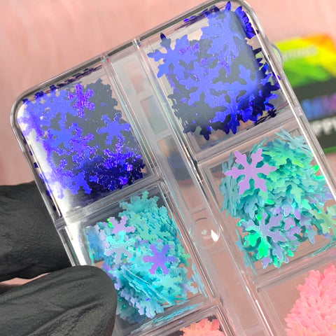 A hand in a black glove holds the Dynamic Nail Supply's Special Iridescent Snowflakes Sequin Glitter against a softly blurred pink background. This clear plastic case is perfect for Christmas nail art, featuring compartments filled with sequins in shades of blue, purple, and teal.