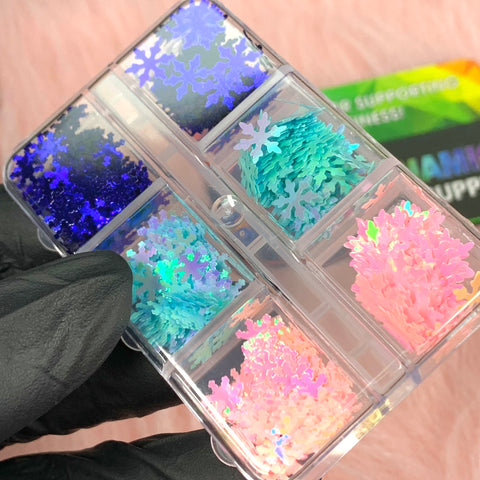 A close-up image features the Dynamic Nail Supply's Special Iridescent Snowflakes Sequin Glitter for Christmas Nail Art Design. The clear plastic container has six compartments filled with shimmering sequins in blue, purple, teal, pink, and white. A gloved hand holds this dazzling assortment against a soft pink background.