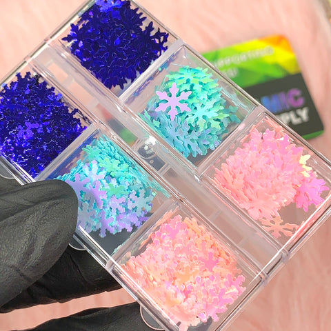 A gloved hand holds a transparent container filled with Special Iridescent Snowflakes Sequin Glitter from Dynamic Nail Supply, showcasing six compartments of glitter in shades from dark blue to pastel pink, green, and blue. In the background on a soft pink surface, a colorful card is partially visible—ideal for Christmas nail art.