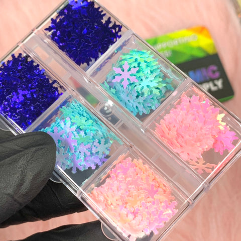 A close-up of a hand wearing a black glove holds a clear plastic container from Dynamic Nail Supply, featuring six compartments filled with the brand's Special Iridescent Snowflakes Sequin Glitter for Christmas Nail Art design. Each compartment contains holographic flakes in vibrant shades of blue, green, pink, and purple, while a holographic product card shimmers in the background.