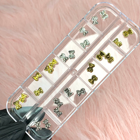A set of 24 silver and gold teddy bear charms for nail art design by Dynamic Nail Supply is neatly stored in a clear plastic organizer featuring two rows of five compartments each. Set against a soft pink, fluffy background, the container is delicately held by someone wearing a black glove.