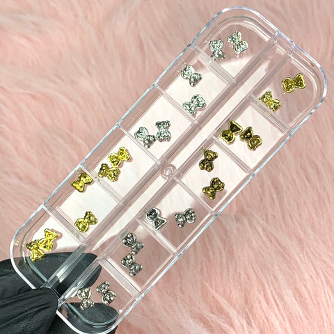 A gloved hand holds a clear plastic box from Dynamic Nail Supply, showcasing small compartments filled with silver and gold teddy bear nail charms. Set against a plush pink background, the box emphasizes the elegant allure of these nail art design adornments.