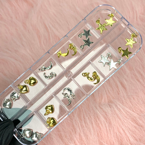 A 24-piece set of silver and gold star, moon, and heart-shaped dangles for nail art design is neatly organized in a transparent container. This Dynamic Nail Supply product is displayed on a pink, fluffy surface, with each charm arranged in separate compartments.