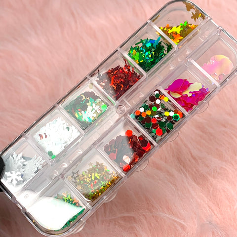 A clear plastic organizer from Dynamic Nail Supply, boasting twelve compartments, rests on a fluffy pink surface. Each section is filled with the Mixed-shapes Christmas Sequin and Glitter for Nail Art Designer, presenting vibrant shades of red, green, white, silver, and iridescent hues in shapes like trees, crosses, snowflakes, and stars for stunning nail art.