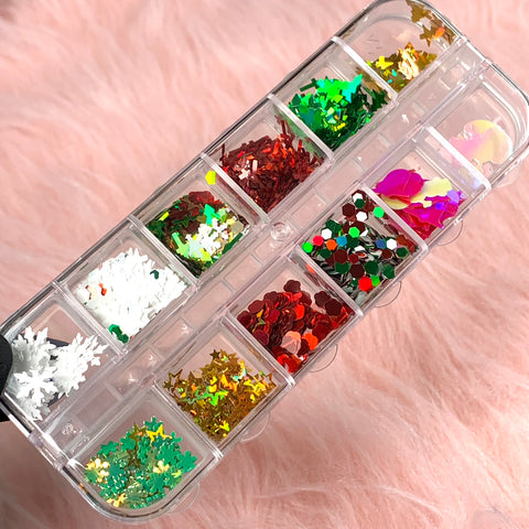 A rectangular plastic container from Dynamic Nail Supply features twelve compartments filled with vibrant Christmas sequin and glitter for nail art. Inside, alongside stars, there are tree, cross, and snowflake shapes in festive reds, greens, golds, and pinks designed to enhance your nail creations. The scene is set against a soft pink, fluffy fabric backdrop.