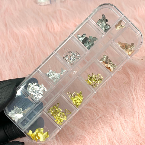 A hand wearing a black glove is holding a clear plastic organizer containing the "3D Butterfly Charms and Sequins set for nail art design" by Dynamic Nail Supply. The organizer features 12 compartments, each filled with shimmering metallic butterfly charms in silver, gold, and rose gold, set against a soft pink, fuzzy background.