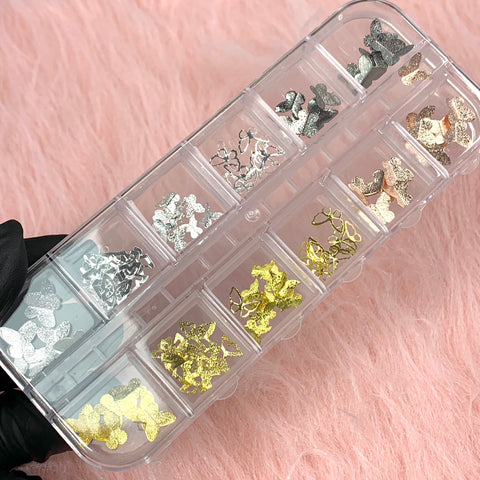 A gloved hand from Dynamic Nail Supply showcases a clear plastic organizer containing the 3D Butterfly Charms and Sequins set for nail art design. The compartments feature silver, gold, and bronze sequins, all beautifully displayed against a soft pink furry fabric backdrop.