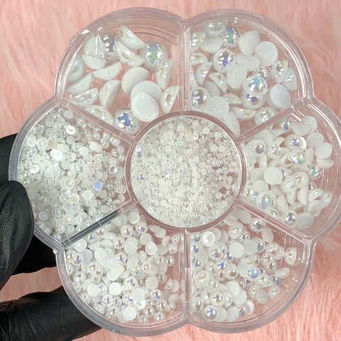 A round, flower-shaped container from Dynamic Nail Supply features an assortment of small, iridescent white beads and gems. A hand in a black glove holds it against a soft pink background. Each compartment contains various shapes, ideal for your next nail art design using Dynamic Nail Supply's White Half Pearl Beads (flat back).