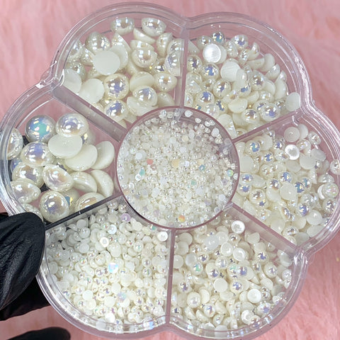 A flower-shaped container filled with an assortment of glossy, matte, and iridescent white half pearl beads for nail art design is beautifully displayed. Held by a hand in a black glove against a soft pink fabric backdrop, this exquisite collection from Dynamic Nail Supply offers a perfect touch for your nail designs.