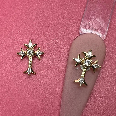 Two cross-shaped charms with rhinestones from Dynamic Nail Supply are displayed on a pink surface. One charm rests on the surface, and the other is attached to a clear backing strip. The pointed ends and symmetrical design of these charms add a delicate and elegant touch to your nail art collection.
