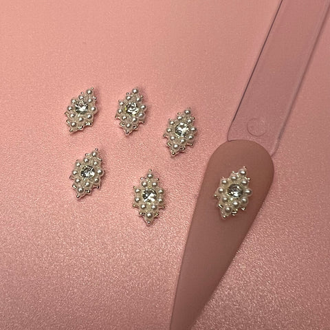 Six rhombus-shaped nail embellishments from Dynamic Nail Supply are arranged on a pink surface, each featuring pearls engraved around a central rhinestone. A nail stand next to them highlights one charm, showcasing its elegant and intricate nail art design.