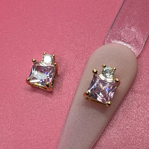 Displayed on a pink background, the 2 pcs 3D Luxury Charm for Nails Art Design from Dynamic Nail Supply exudes elegance with its gold prong setting. Each charm features a square iridescent pink rhinestone topped with a smaller round gem. One piece is artfully positioned on a beige nail tip, showcasing its stylish design and perfect fit for nail art enthusiasts.