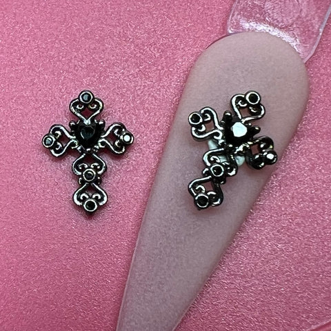 On a pink surface lie two charms from Dynamic Nail Supply, known as the "2 pcs 3D Black Crosses Charm for Nails Art Design." These decorative metallic nail charms feature intricate heart-shaped patterns and are adorned with black rhinestones at their center. One charm is elegantly attached to a nail tip display, adding sophistication to the design.