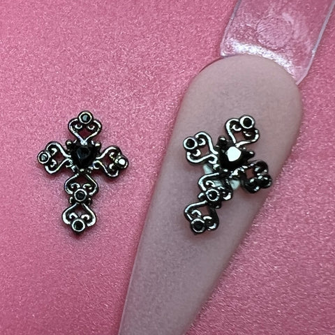 Two decorative black cross-shaped charms from Dynamic Nail Supply sit atop a pink, textured surface. One charm adorns a beige nail tip, highlighting the 2 pcs 3D Black Crosses Charm for Nails Art Design with intricate loops and a central black rhinestone. This nail art design radiates elegance and sophistication with its exquisite details.