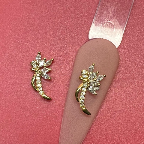 Two gold and diamond stud earrings sit on a pink surface, each adorned with a floral design featuring a curved stem of gold and clear marquise-cut stones as petals. Nearby, an elegant 2-piece 3D Metal Charm for Nails Art Design from Dynamic Nail Supply complements the scene, showcasing delicate sophistication.