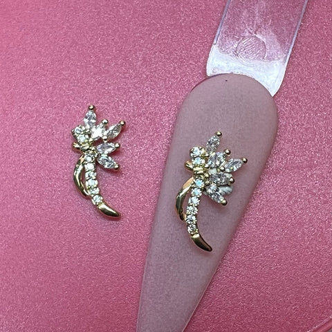 Gold and crystal earrings shaped like delicate flowers, with marquise and round-cut stone petals, are showcased against a soft pink background that accentuates their intricate design. They pair beautifully with the "2 pcs 3D Metal Charm for Nails Art Design" from Dynamic Nail Supply, adding a touch of sparkle and elegance to your look.