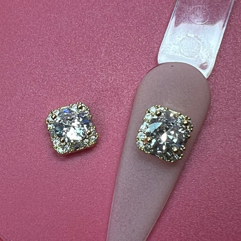 A close-up shows two cushion-cut diamond earrings set in gold on a pink textured background, each featuring surrounding smaller diamonds to enhance their brilliance. Beside them sits a luxury nails art design from Dynamic Nail Supply's 2 pcs Clear Crystal Square Charm for Luxury Nails Art Design (Crystal Rhinestone Engraved), perfectly complementing the sparkle of the jewelry.