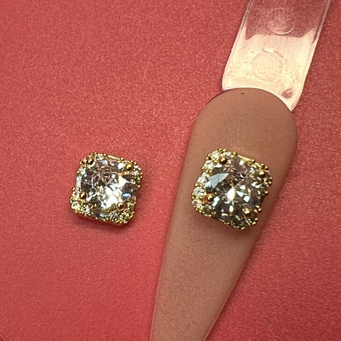A close-up shot showcases the two-piece Clear Crystal Square Charm set from Dynamic Nail Supply, designed for luxury nails art. The crystal rhinestone engraved charms are elegantly placed on a smooth pink surface, with intricately detailed edges enhancing their brilliance. One charm rests atop a pink curved nail swatch, seamlessly uniting luxury nail art with an elegant presentation.