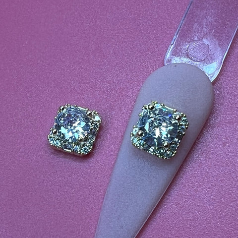 Close-up of two square crystal charms with gold settings on a pink, textured surface. Each charm, from Dynamic Nail Supply's 2 pcs Clear Crystal Square Charm for Luxury Nails Art Design collection, resembles clear crystal square magic and showcases a large central crystal rhinestone surrounded by smaller embellishments. One charm is displayed on a tapered pink stand, enhancing the presentation's elegance.