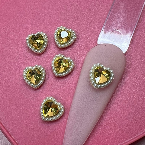 Five golden heart-shaped charms featuring engraved pearl edges from Dynamic Nail Supply's collection are stylishly placed on a pink surface. One glimmering heart-shaped charm is elegantly attached to a matte mauve almond-shaped nail tip, producing a striking contrast.
