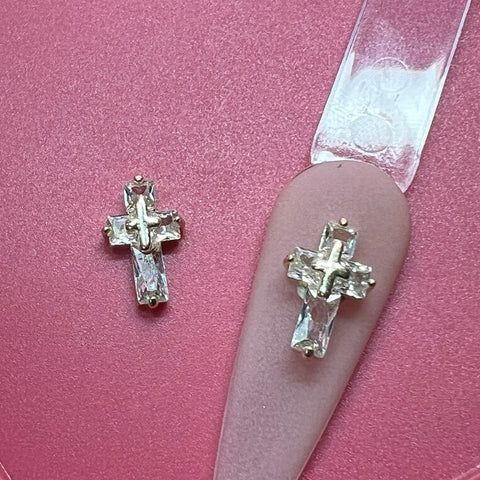 A close-up of the 2 pcs 3D Crucify Charm for Nails Art Design from Dynamic Nail Supply on a pink background. One charm embellishes a pink nail tip attached to a clear strip, featuring an intricate design. Both crosses sparkle with silver-toned settings, offering an elegant and decorative appearance.