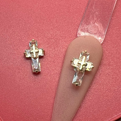 Two cross-shaped earring charms from Dynamic Nail Supply, featuring a gold finish with sparkling crystal accents, rest on a soft matte pink surface. These 3D Crucify Charms for nails art design are intricately crafted, with rhinestones embedded in each cross, adding an elegant touch. One charm is positioned on the edge of a beige nails art design palette.
