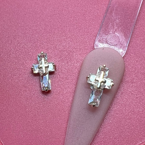 Two Dynamic Nail Supply 3D Crucify Charms for Nails Art Design, adorned with clear rhinestones, gleam on a shiny pink surface. One charm elegantly rests on a nail art design display, showcasing its potential style. The gold metal accents provide a stunning contrast against the pink backdrop, highlighting the intricate craftsmanship of these charms.