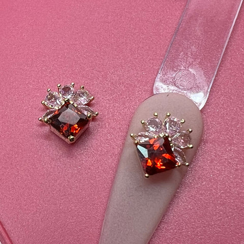 On a pink surface lie two luxurious gemstone earrings, each showcasing a large red rhinestone with a square shape at the center. Above it, smaller, clear teardrop-shaped gems are elegantly arranged. One of these dazzling earrings is positioned on a 2 pcs Luxury 3D Charm for Nails Art Design tool by Dynamic Nail Supply, emphasizing its ornamental charm and luxury appeal.